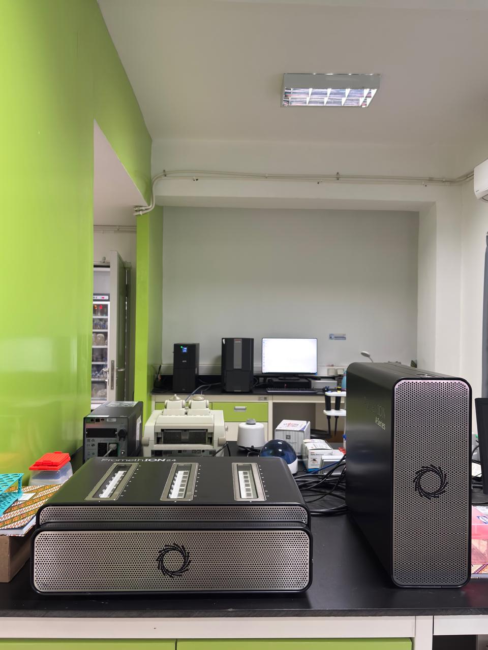 Perangkat sequencing DNA PromethION A-Series dari Oxford Nanopore Technologies yang terdiri dari unit pemroses utama berwarna hitam di sebelah kanan, storage dock dengan sistem ventilasi perforated di sebelah kiri, dan tiga flow cell modules dapat dilepas pasang yang menampilkan display dan indikator status biru untuk analisis sekuens genom real-time